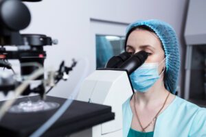 A fertility lab technician wearing a mask and cap looks into a microscope.