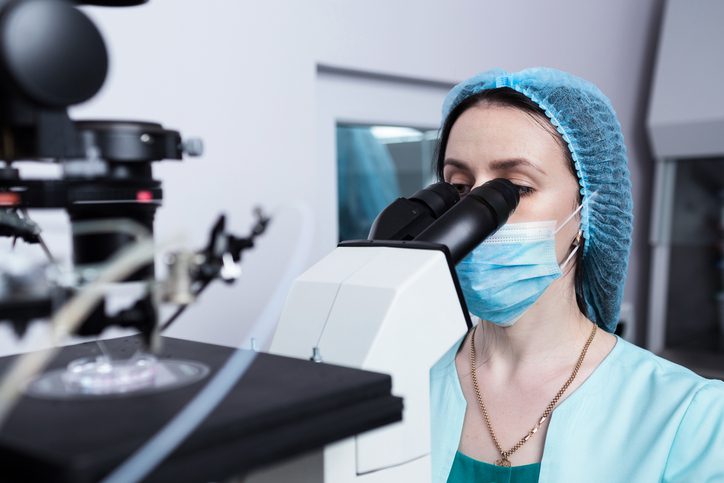 A fertility lab technician wearing a mask and cap looks into a microscope.