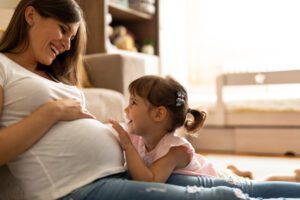 Smiling pregnant woman sits on the floor as a young girl joyfully touches her belly.