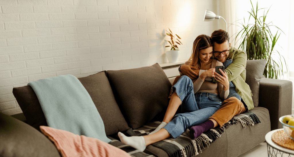 A joyful couple cuddles on a couch, looking at a phone, symbolizing hope and excitement as they plan their parenthood journey together.