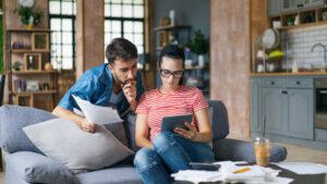 Couple calculating bills at home using tablet and calculator. Young couple working on computer while calculating finances sitting on couch. Young man with wife at home analyzing their finance with documents.