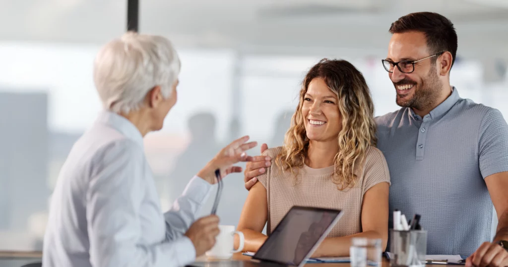 A smiling couple engages in a warm conversation with a professional, expressing optimism and trust. The setting suggests a consultation, where guidance and support play a key role in their journey.