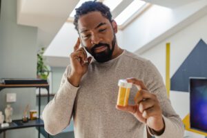 African American man is at home. He is communicating with the doctor via mobile phone while holding a bottle of medicine in his hand.