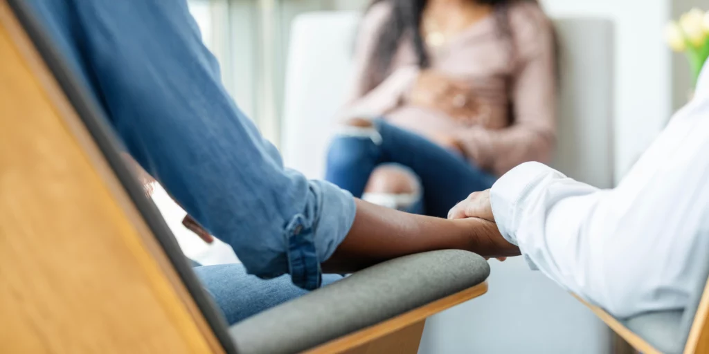 Two people hold hands in the foreground while a pregnant woman sits in the background.