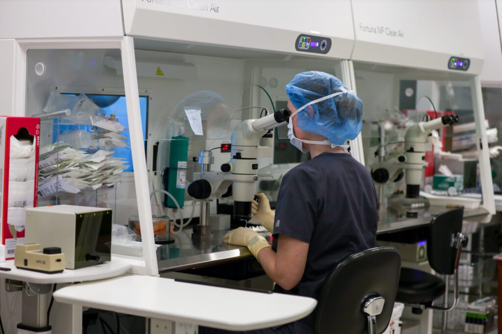A laboratory technician in a fertility clinic examines samples under a high-powered microscope inside a sterile environment, ensuring precise evaluation for in vitro fertilization (IVF) procedures.