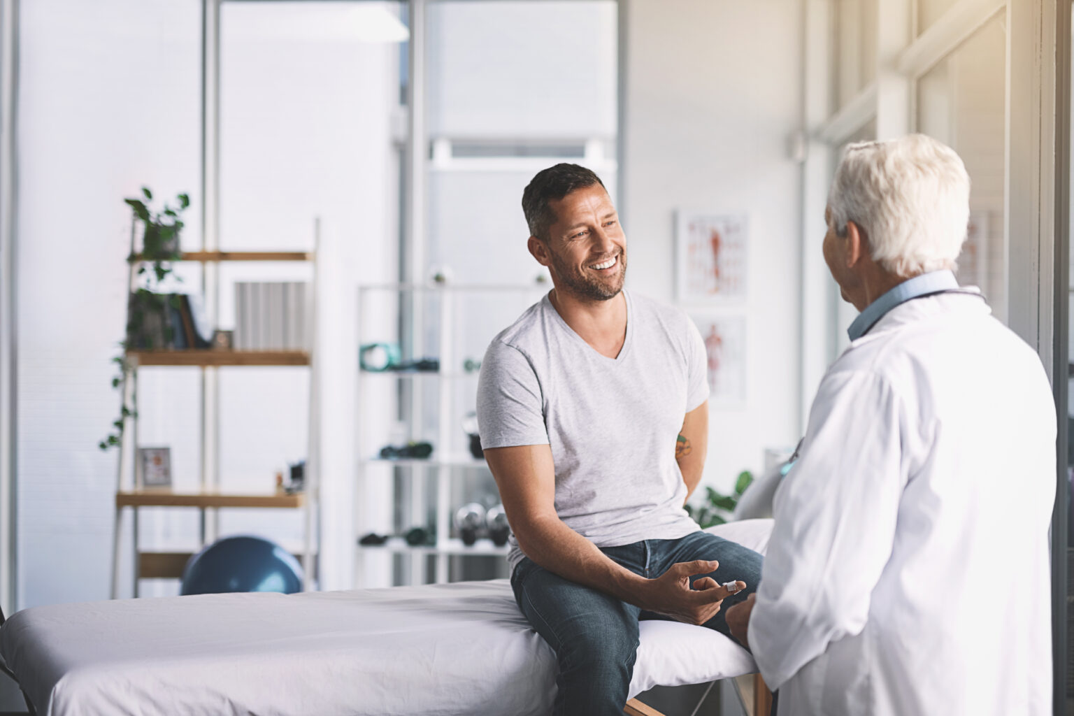 A man sits on an exam table, smiling while talking to a doctor, possibly discussing fertility treatment