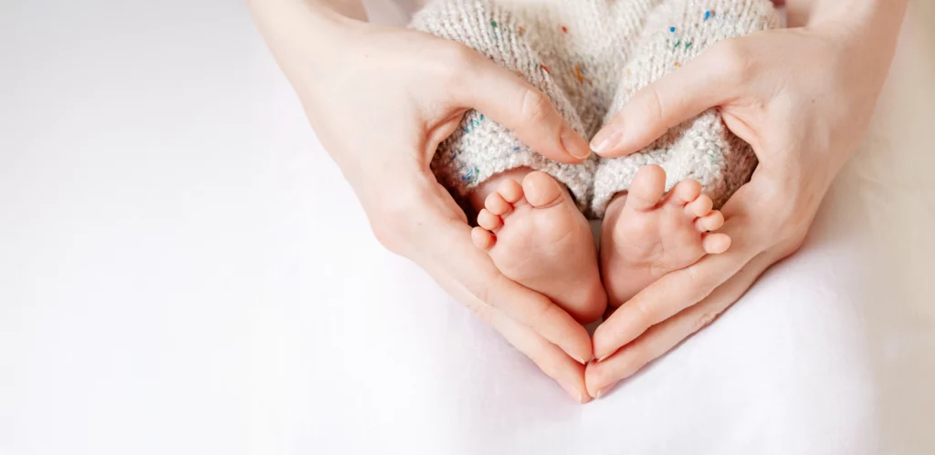 A loving parent cradles their newborn’s tiny feet in their hands, forming a heart shape as a symbol of warmth, care, and the deep bond between parent and child.