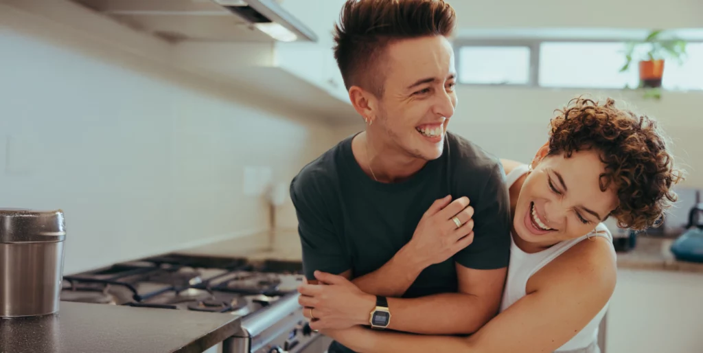 A joyful couple embraces in a kitchen, laughing together. The image conveys love, warmth, and connection in a relaxed home setting.