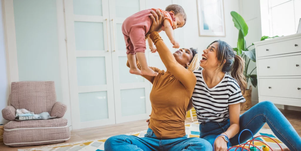 A joyful lesbian couple plays with their baby in a bright, cozy home. The image reflects love, family, and LGBTQ+ parenting.