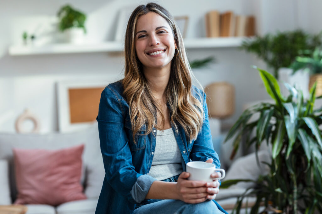 Beautiful young woman drinking a cup of coffee while sitting on couch in living room at home.