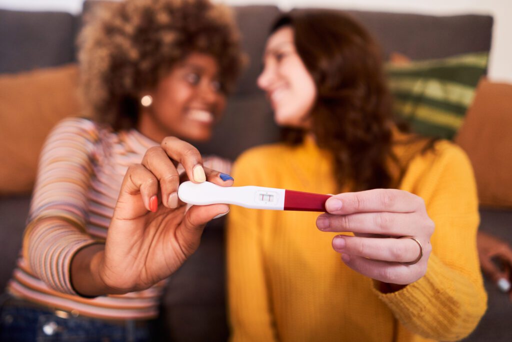 Expecting lesbian multicultural couple at home showing to camera the positive pregnancy test with focus on it