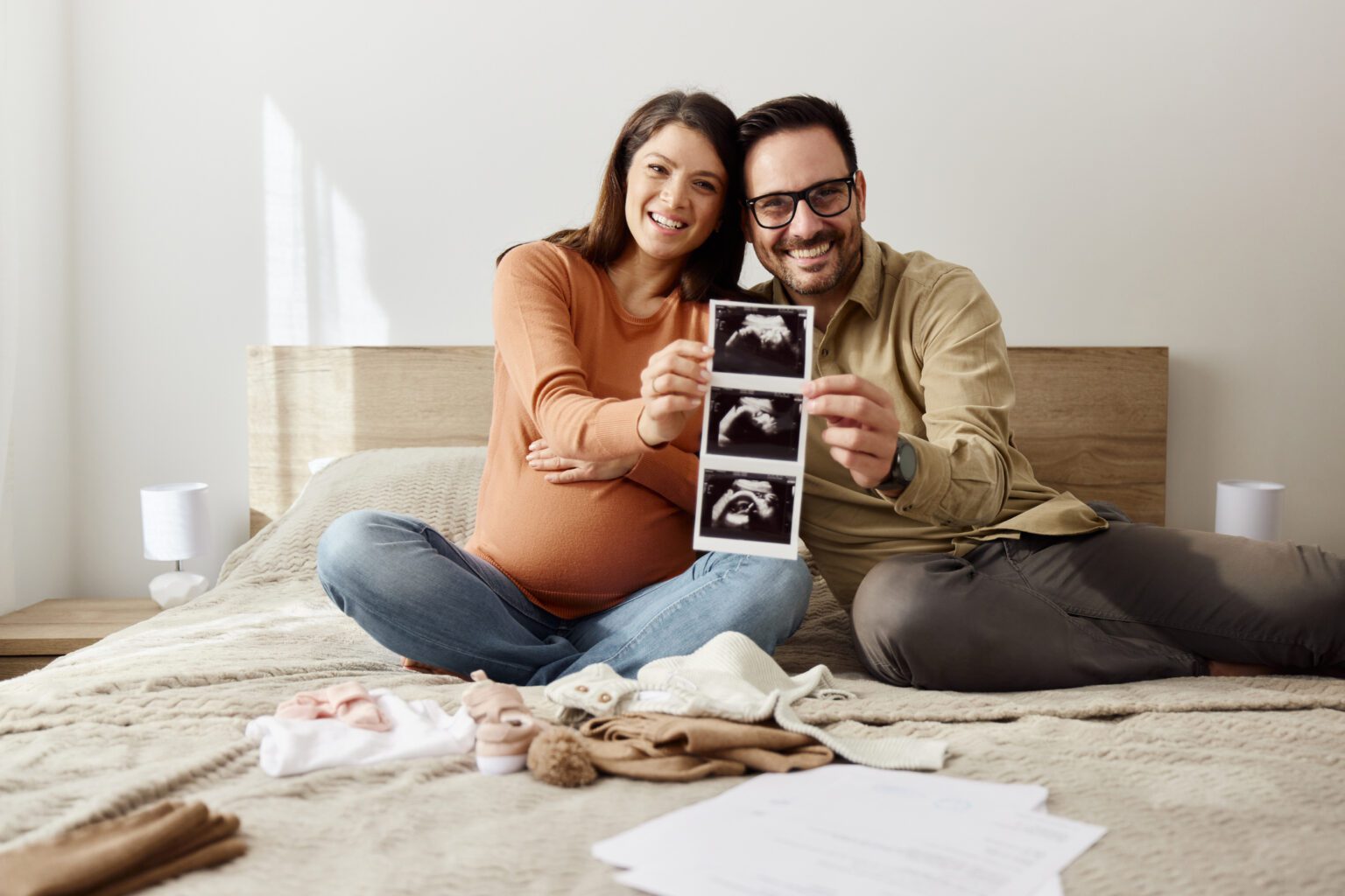 Happy pregnant couple showing medical scan of their baby at home and looking at camera. Copy space.