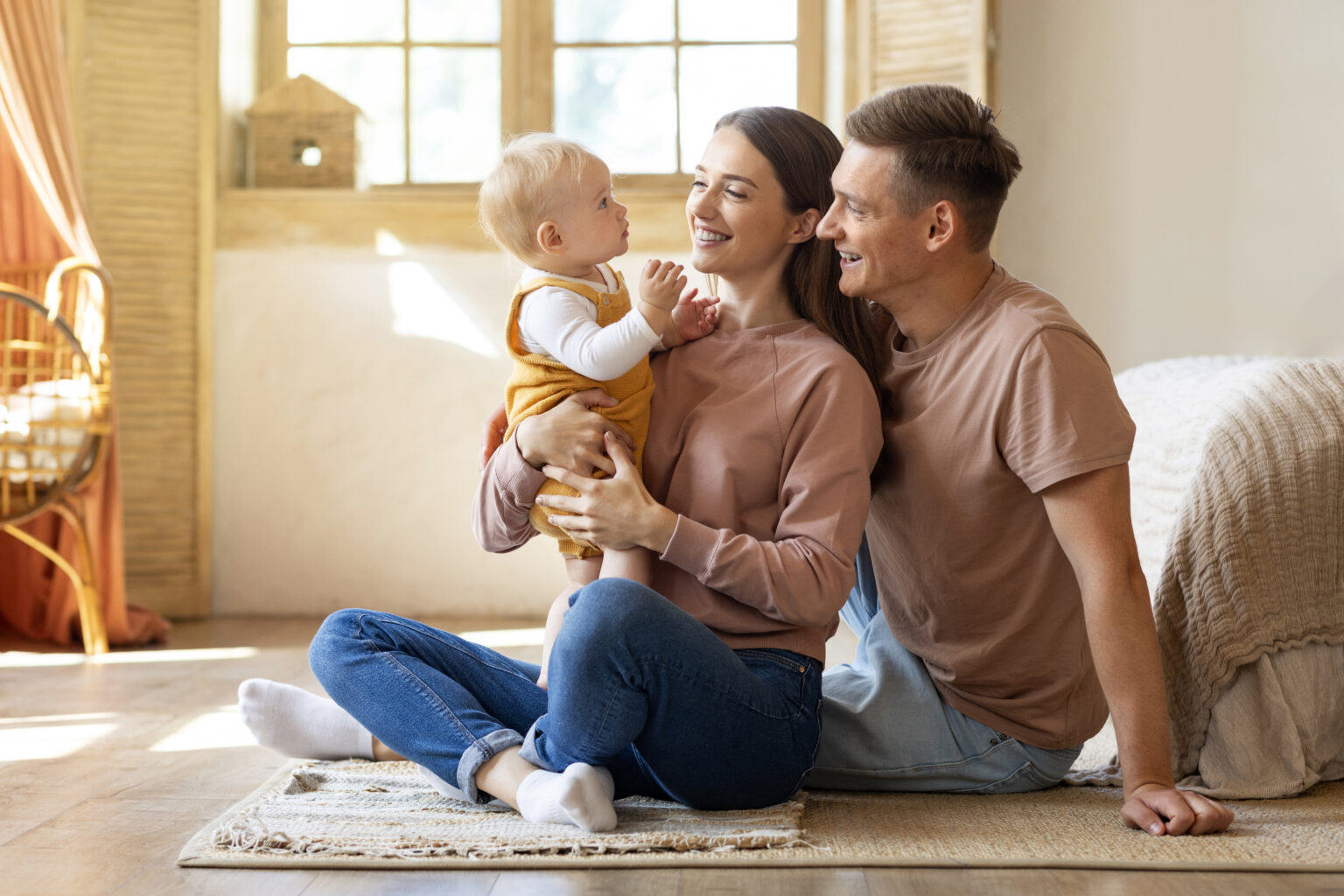A happy family sits on the floor, smiling as they interact with their baby in a warm, sunlit home, sharing a loving moment together.