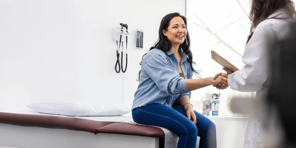 A smiling patient in a medical exam room shakes hands with a doctor, indicating a friendly consultation or appointment.