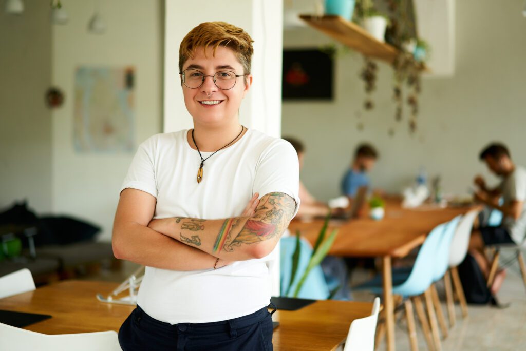 Confident transgender male employee smiling in office space, arms crossed, diverse team working in background.