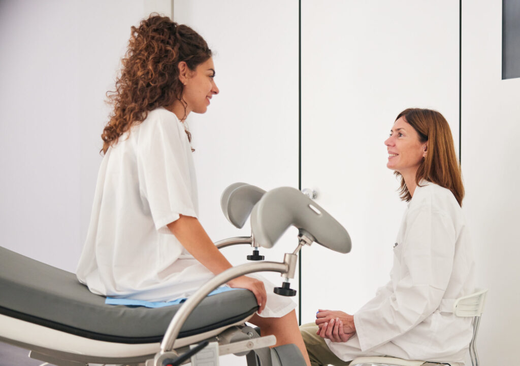 The young woman, dressed in a hospital gown, sits on the stretcher as the mature doctor in a white lab coat sits on a stool, and they engage in discussion, looking at each other.