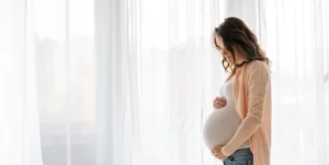 Pregnant woman in casual clothes gently holds her belly, standing by a bright window with white curtains.