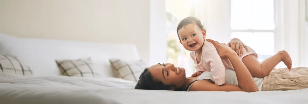 Mother lying on a bed, smiling while holding her happy baby in a bright, cozy room.