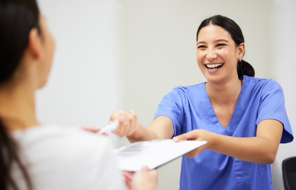 A smiling nurse in blue scrubs hands a clipboard and pen to a patient, likely for form completion in a medical setting.