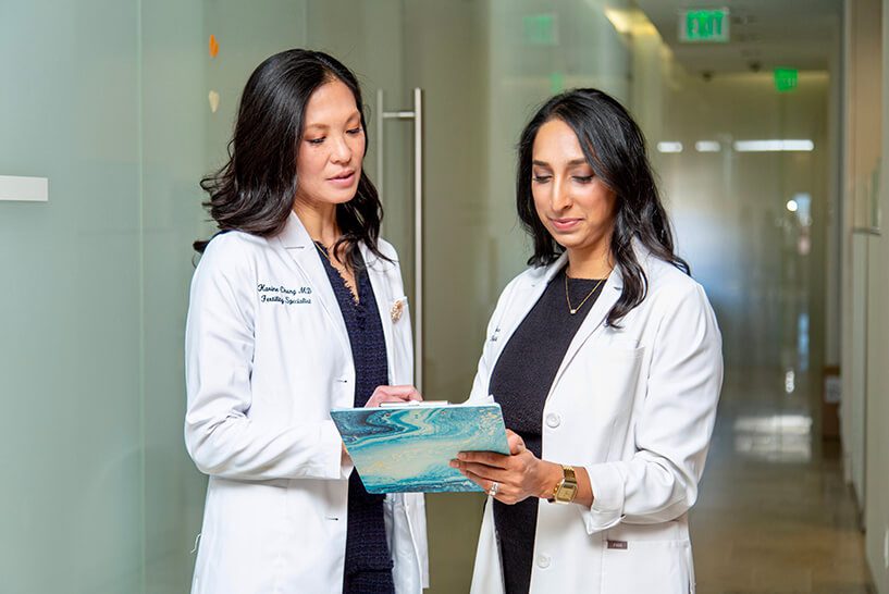 Two female doctors in white coats review a document in a modern medical office hallway, engaged in discussion.