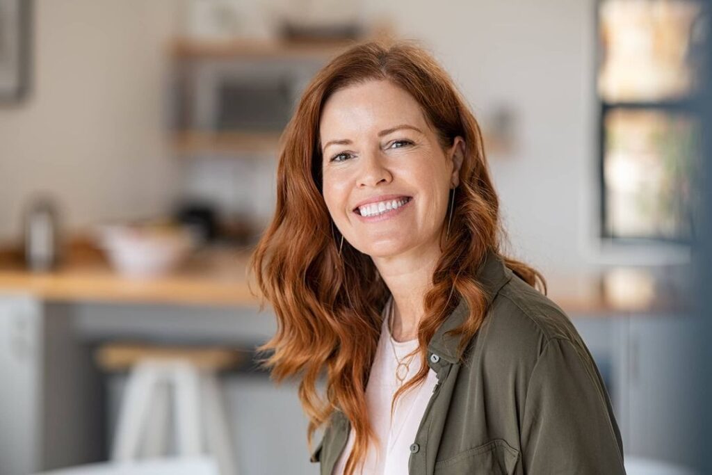 Smiling middle-aged woman with long red hair in a casual button-down shirt, standing in a warmly lit kitchen.