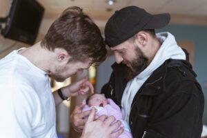 Two Men Holding Newborn Baby Together At Hospital