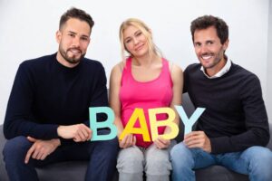 Two men and a pregnant woman sitting on a couch holding colorful letters that spell “BABY,” representing a surrogacy arrangement or LGBTQ+ family building.