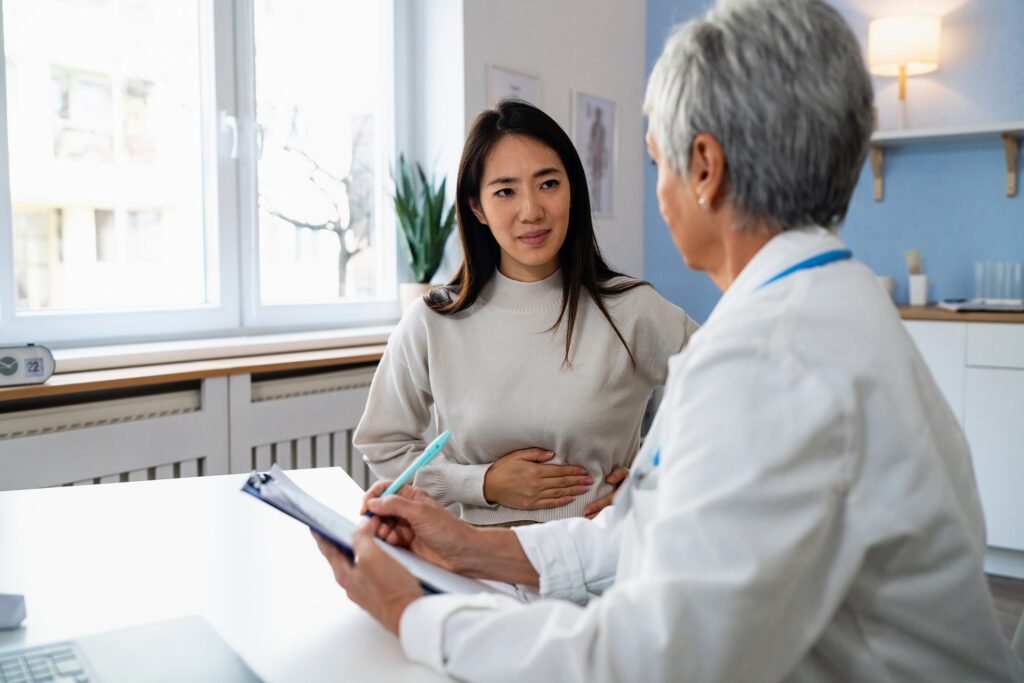 Female patient Japanese ethnicity describes stomach ache symptoms, to senior Caucasian female doctor