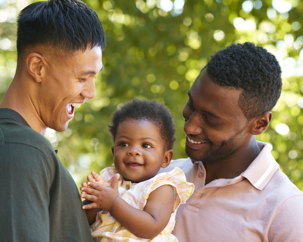 Loving Same Sex Male Couple Or Friends Cuddling Baby Outdoors In Garden Or Countryside Together