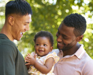 Loving Same Sex Male Couple Or Friends Cuddling Baby Outdoors In Garden Or Countryside Together