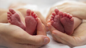 Mother and father’s hands cradling twin babies’ feet on a pale background